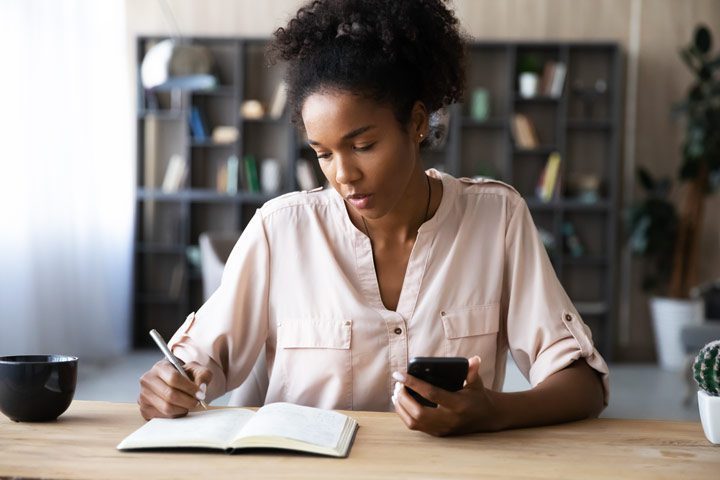 pretty young Black woman making notes in a notebook while holding her cell phone - residential